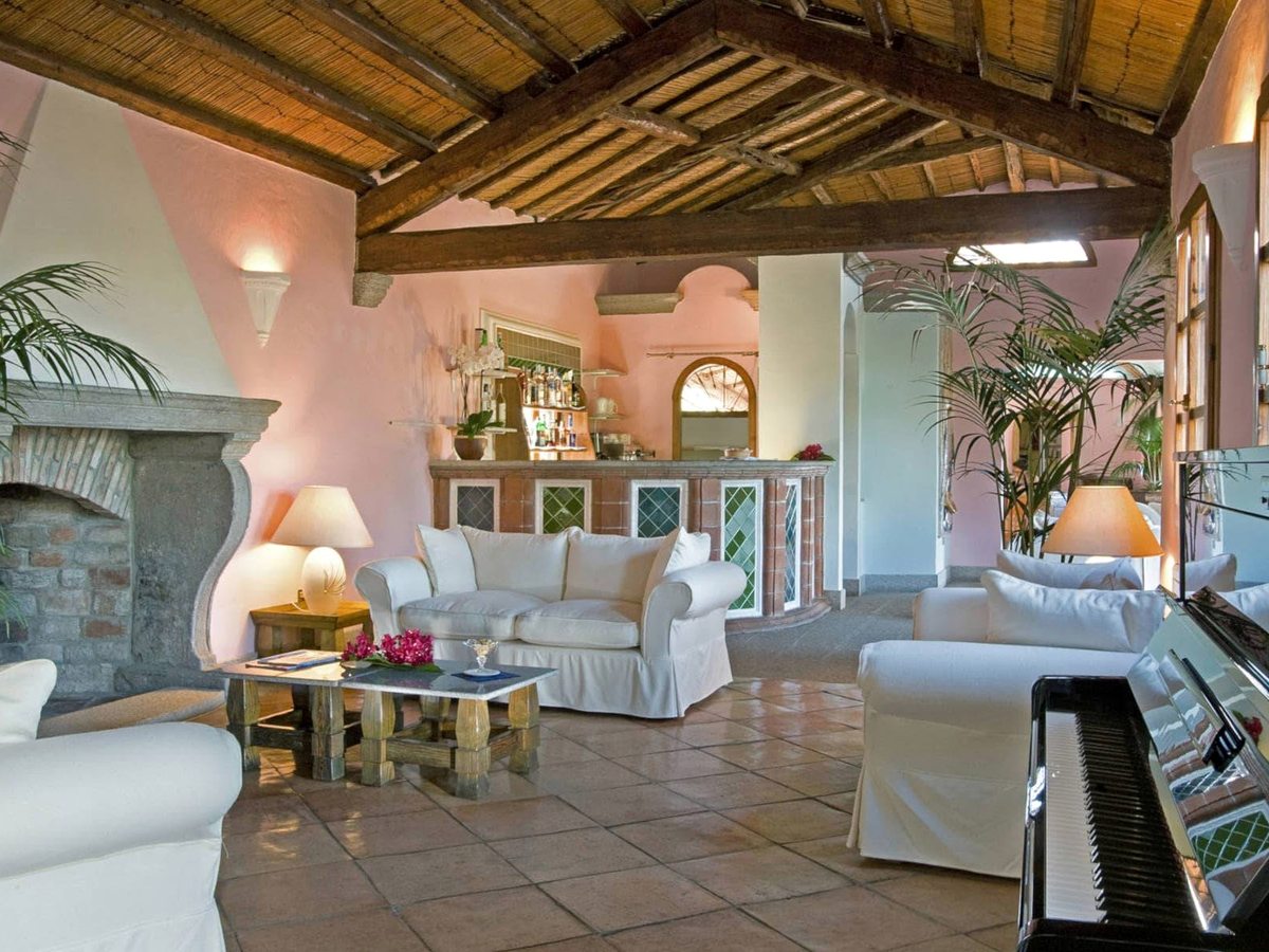 An open lounge with white sofas, a stone fireplace, a tiled bar and a piano beneath a pitched cane-and-timber ceiling.
