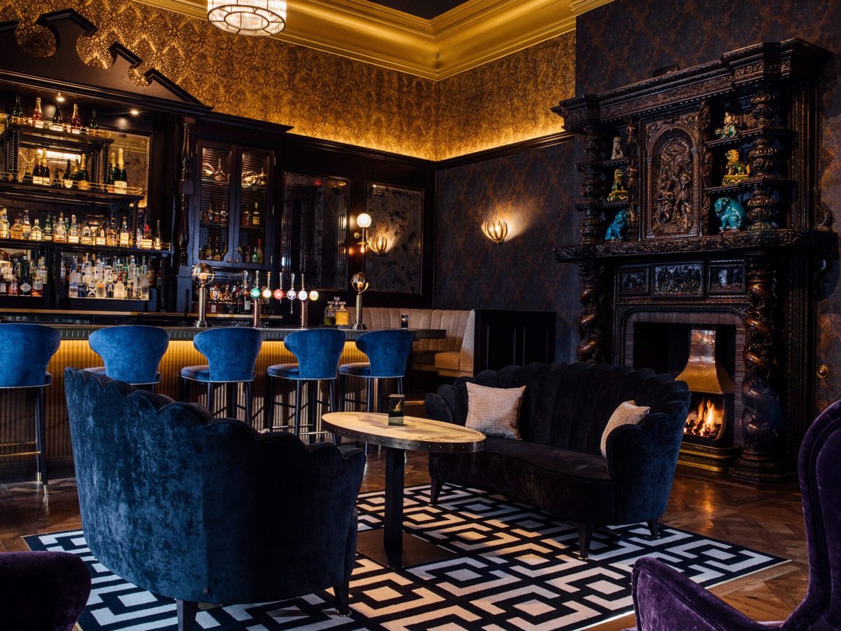 A traditional bar with blue velvet seating, a carved fireplace with a lit fire and a patterned rug.