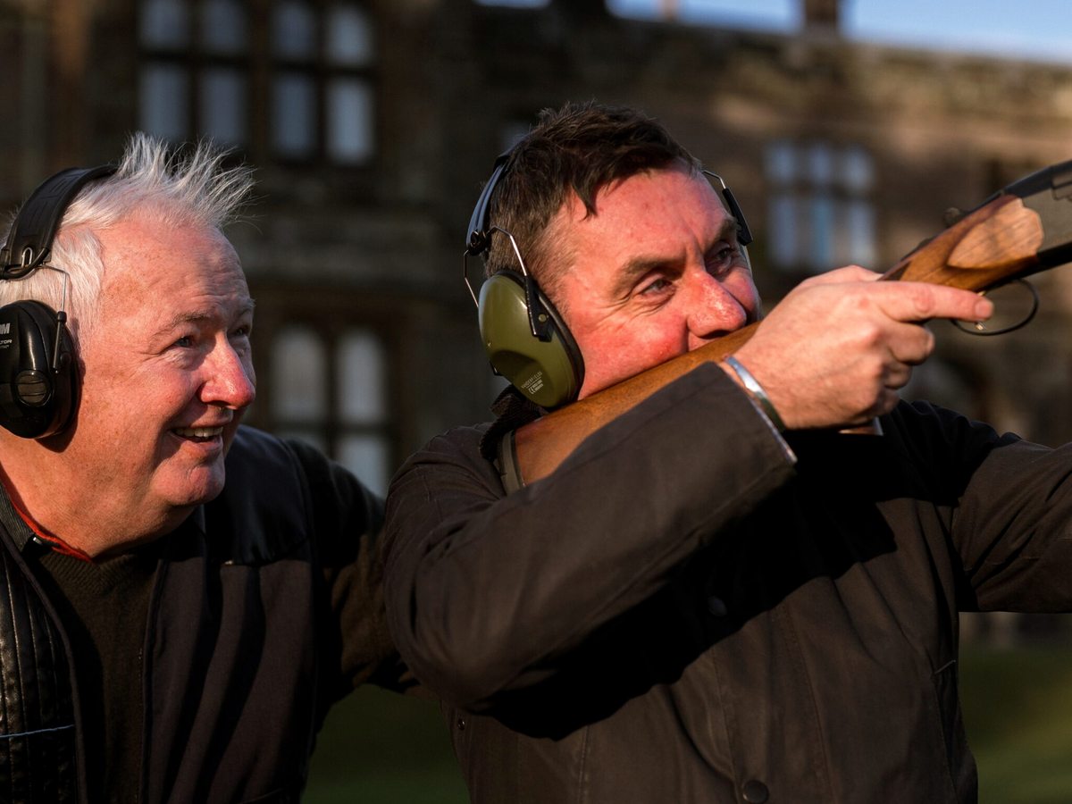 Two men in ear defenders, one shouldering a shotgun, on the estate lawn.