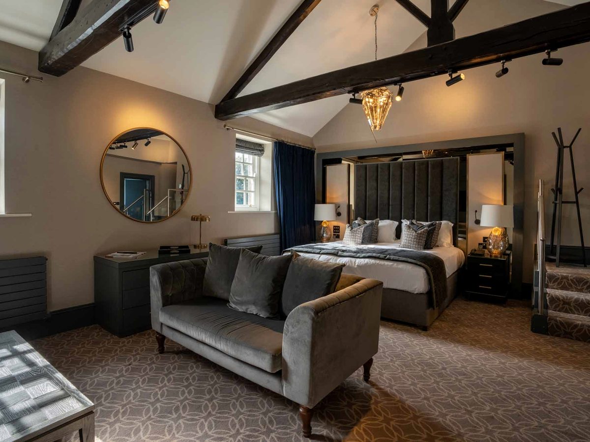 A suite bedroom with a large bed, sofa, round mirror and exposed timber roof beams.