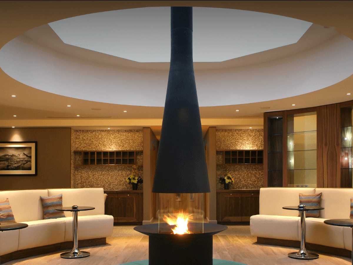 A circular spa lounge with curved cream sofas around a suspended conical fireplace under a ceiling oculus.