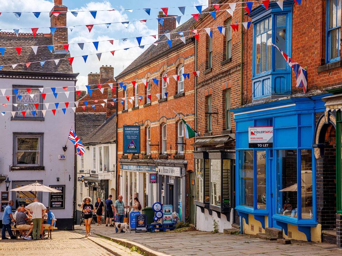 A sloping street of brick shopfronts, one painted blue, with bunting overhead and cafe tables on the pavement.