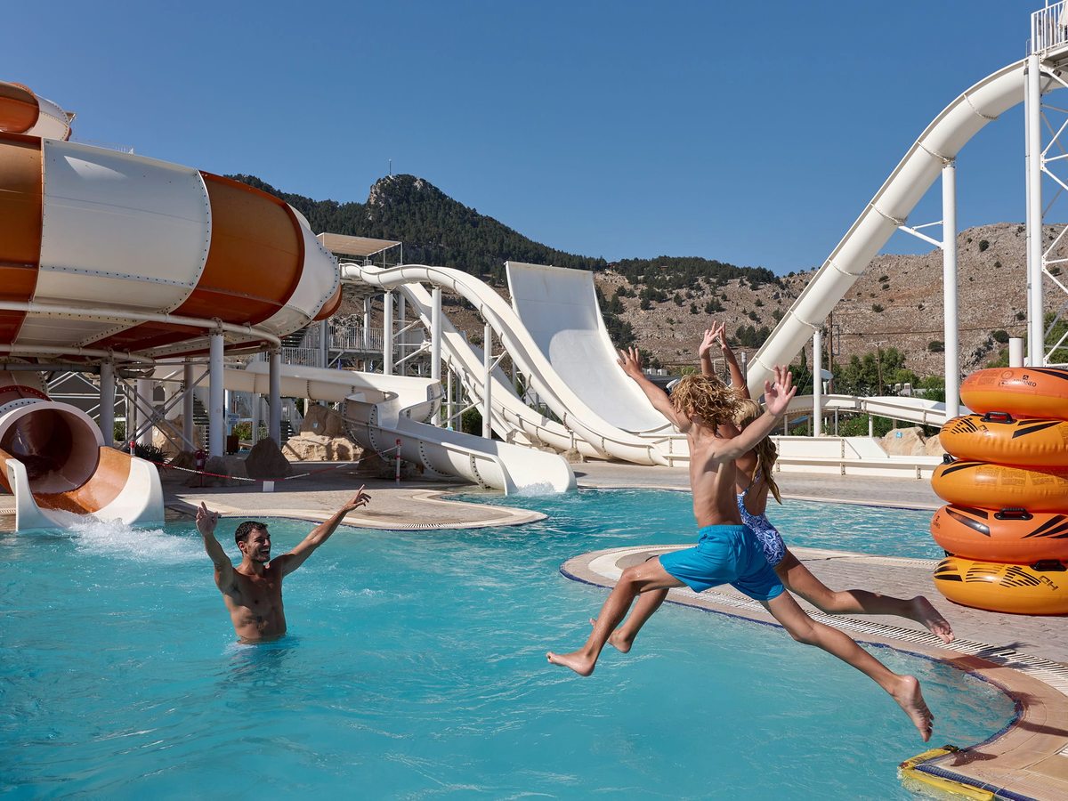 Orange and white tube slides curving down to a splash pool, with a child leaping in and the bare slopes of Tsambika Mountain behind.