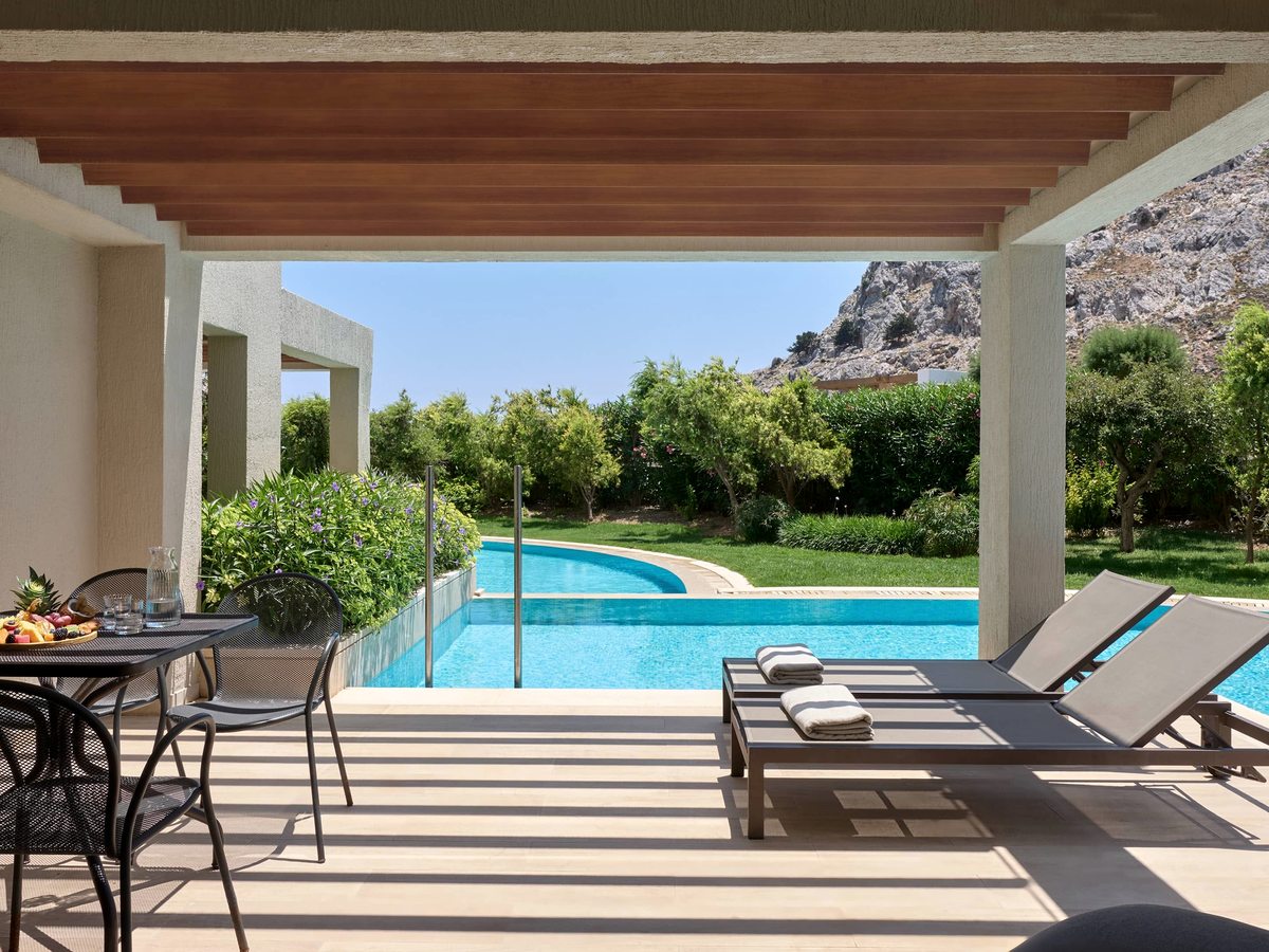 A covered room terrace with sun loungers and a table of fruit, opening onto a private stretch of pool with the rocky mountain behind.