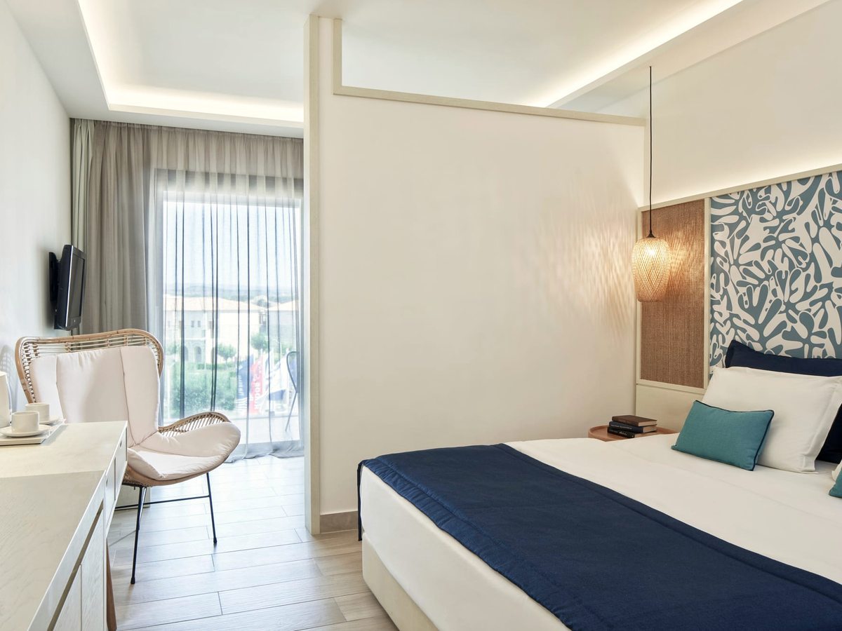 A bright bedroom with a double bed dressed in white and navy, a patterned headboard and a window to the side.