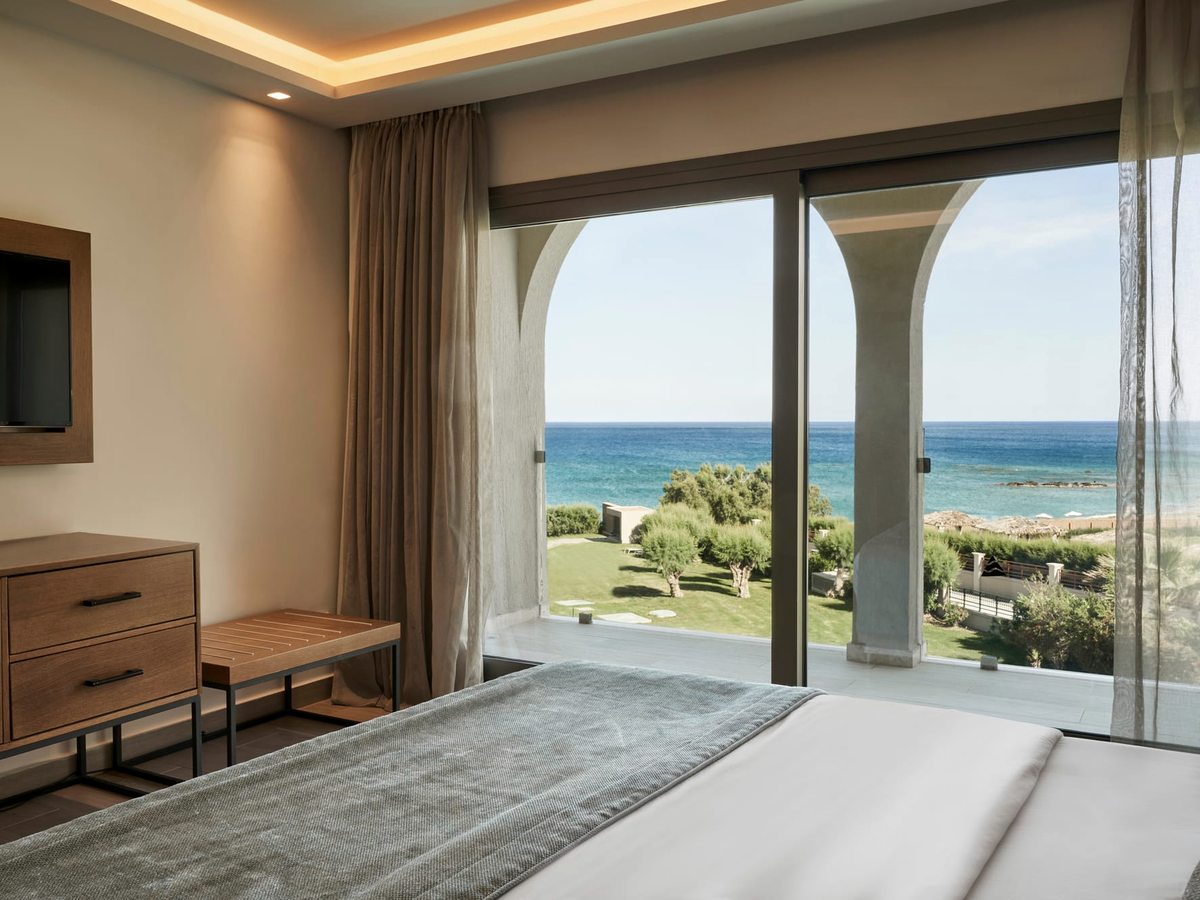 A bedroom with sliding glass doors framing a sea view, a wooden dresser and a wall-mounted television.