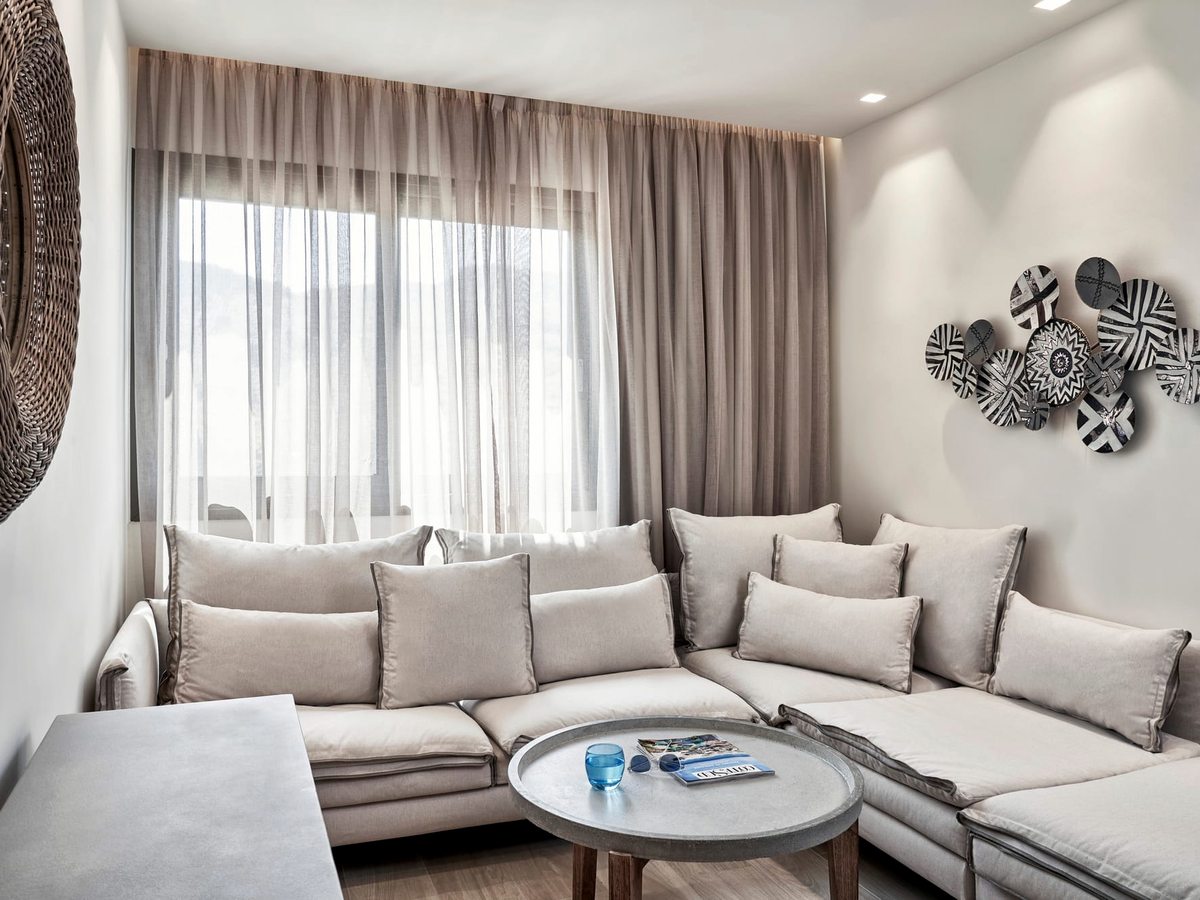 A suite living room with a large L-shaped grey sofa, cushions and a round coffee table beside a curtained window.