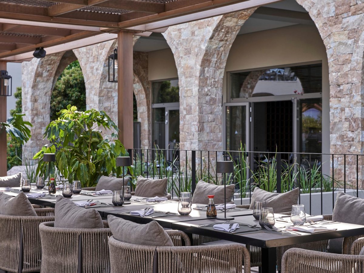 An outdoor restaurant terrace set with dark tables and grey cushioned chairs beneath a timber pergola and tall stone archways.