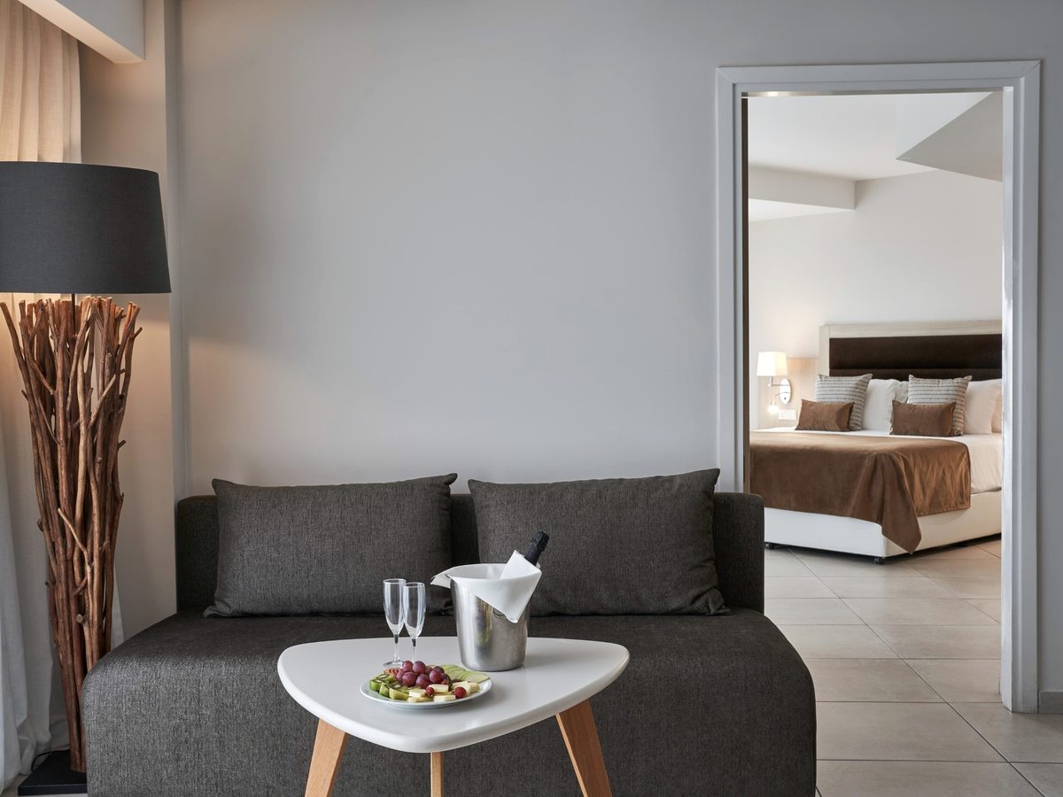 Suite with a dark sofa and coffee table in the sitting room, a wide internal doorway leading through to the double bedroom, with welcome fruit and sparkling wine set out.