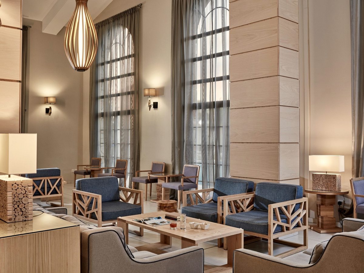 Double-height lobby lounge with blue armchairs and sofas, coffee tables and pendant lights below tall arched windows with sheer curtains.