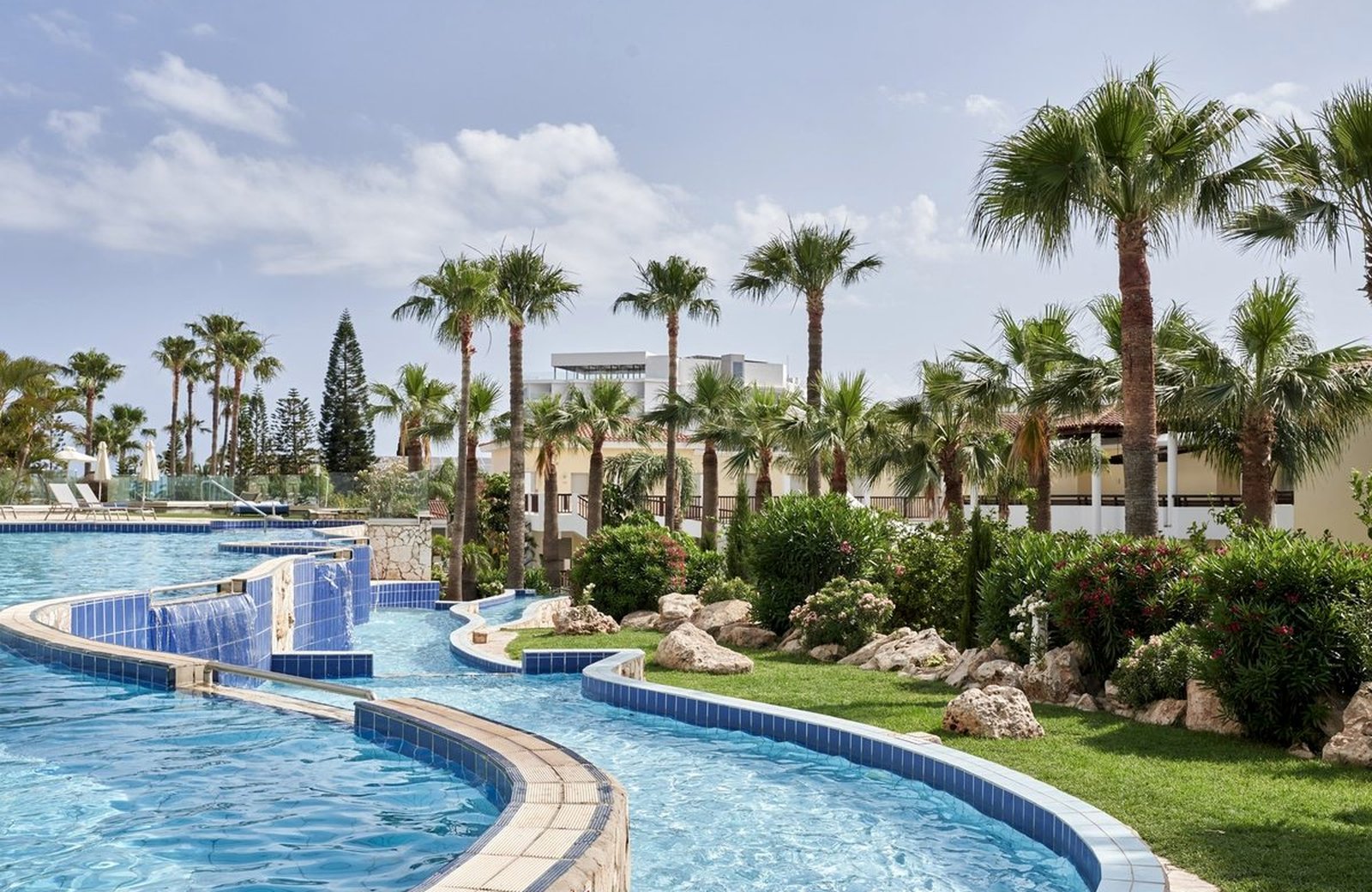 The free-form lagoon pool at Atlantica Aeneas, a waterfall spilling between its levels past tall palms and planted gardens under a clear Cyprus sky.
