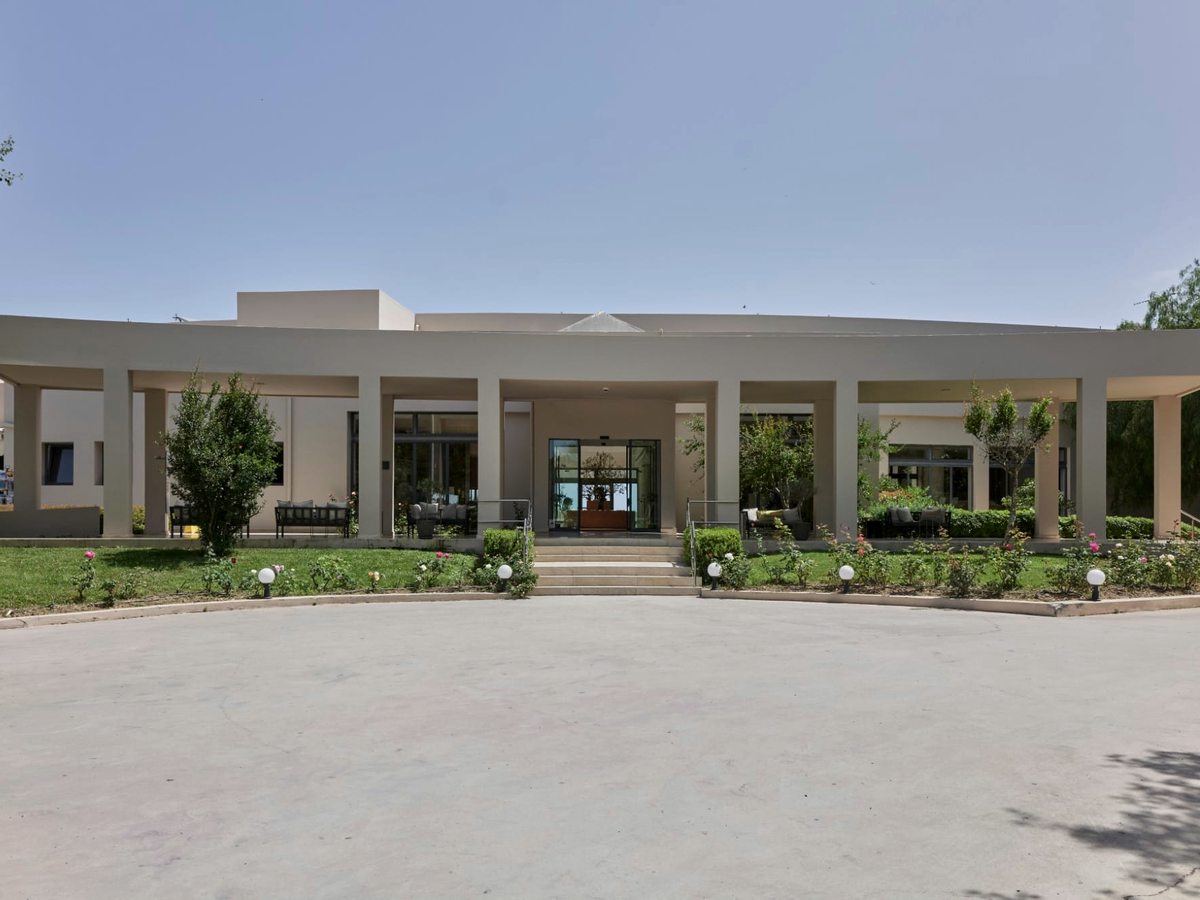 Single-storey reception building with a colonnaded portico, glass entrance doors reached by low steps, rose beds and a wide paved forecourt under a clear sky.