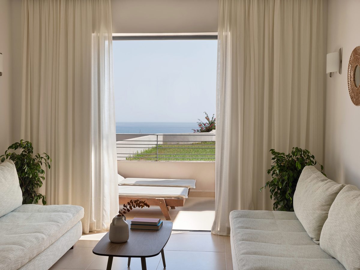 Suite sitting room with a pale sofa and day bed, a low round table and open curtains onto a terrace with a distant sea view.