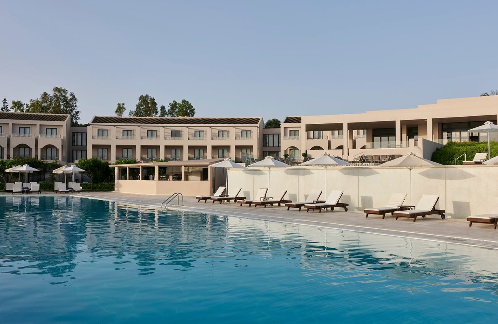 A rectangular outdoor pool with sun loungers and white parasols along the paved deck, the resort's guest blocks and olive trees rising behind under a clear sky.