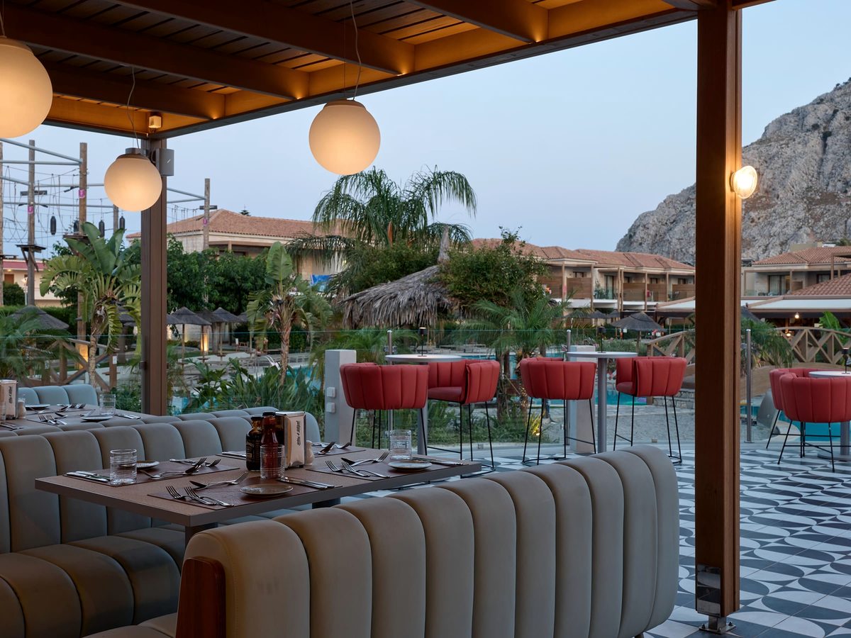Open-air covered restaurant terrace with striped banquette seating, red bar stools and globe pendant lights beside the pool and gardens.