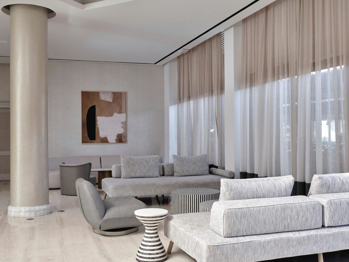 Indoor lounge with pale grey sofas and armchairs, round side tables, tall columns and floor-to-ceiling curtains on a light stone floor.