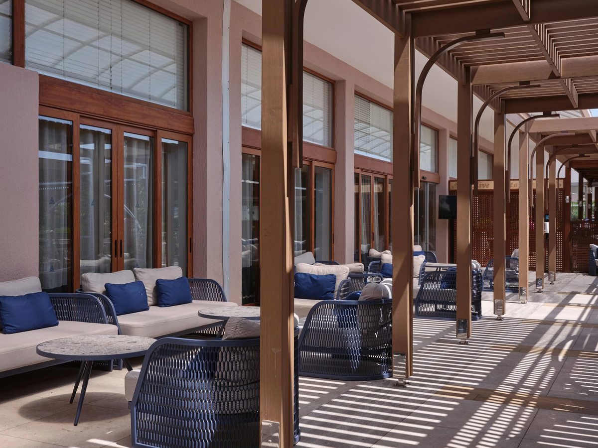 Covered terrace lined with cushioned sofas and woven armchairs, timber columns and a slatted pergola along a tiled walkway.