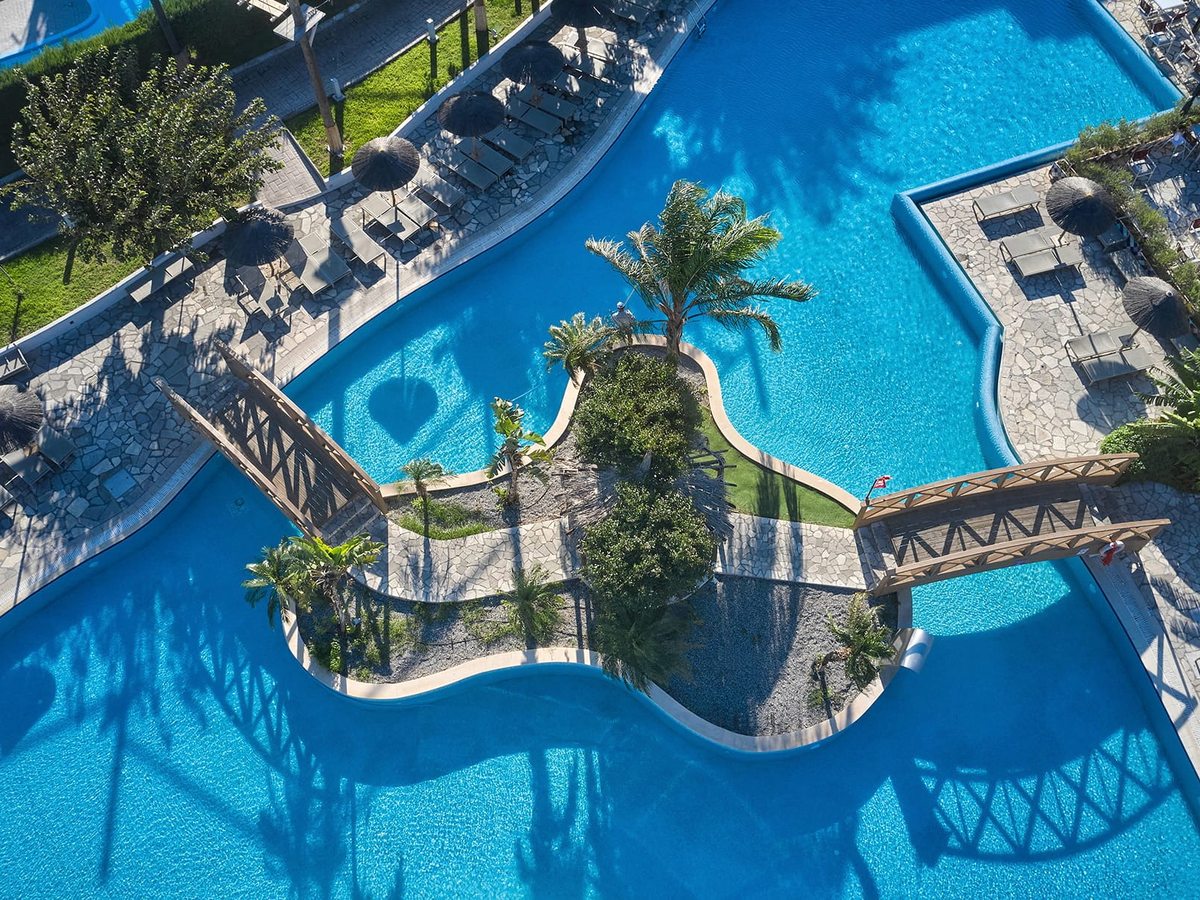 Aerial view of a curving blue pool with two wooden footbridges to a central planted island of palms, loungers and parasols around the edge.