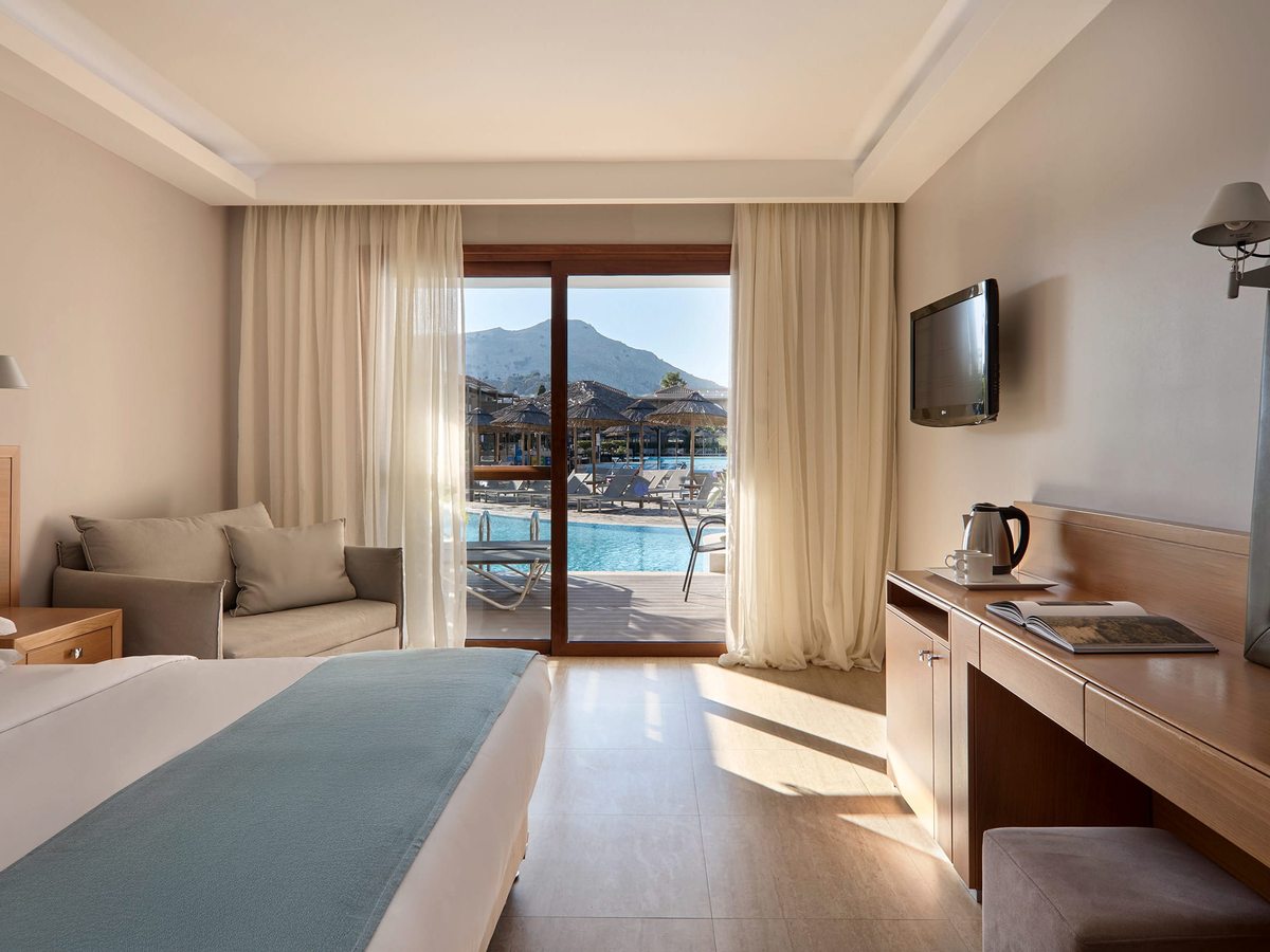 Room with a large bed, a cream sofa and a desk with a kettle, sliding doors to a balcony overlooking the pool and mountains.