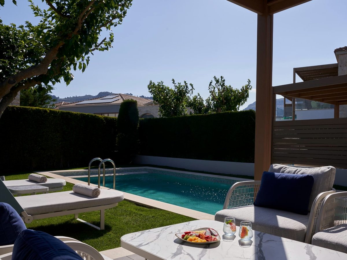 Covered suite terrace with cushioned seating and a fruit plate beside a private rectangular pool, sun loungers and a hedged garden.