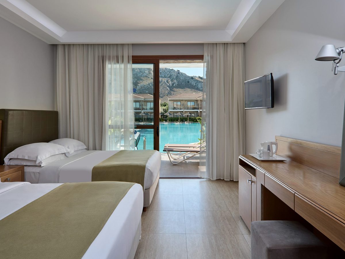 Room with two single beds, a wall-mounted TV and a long desk, sliding doors to a balcony over the pool with mountains beyond.