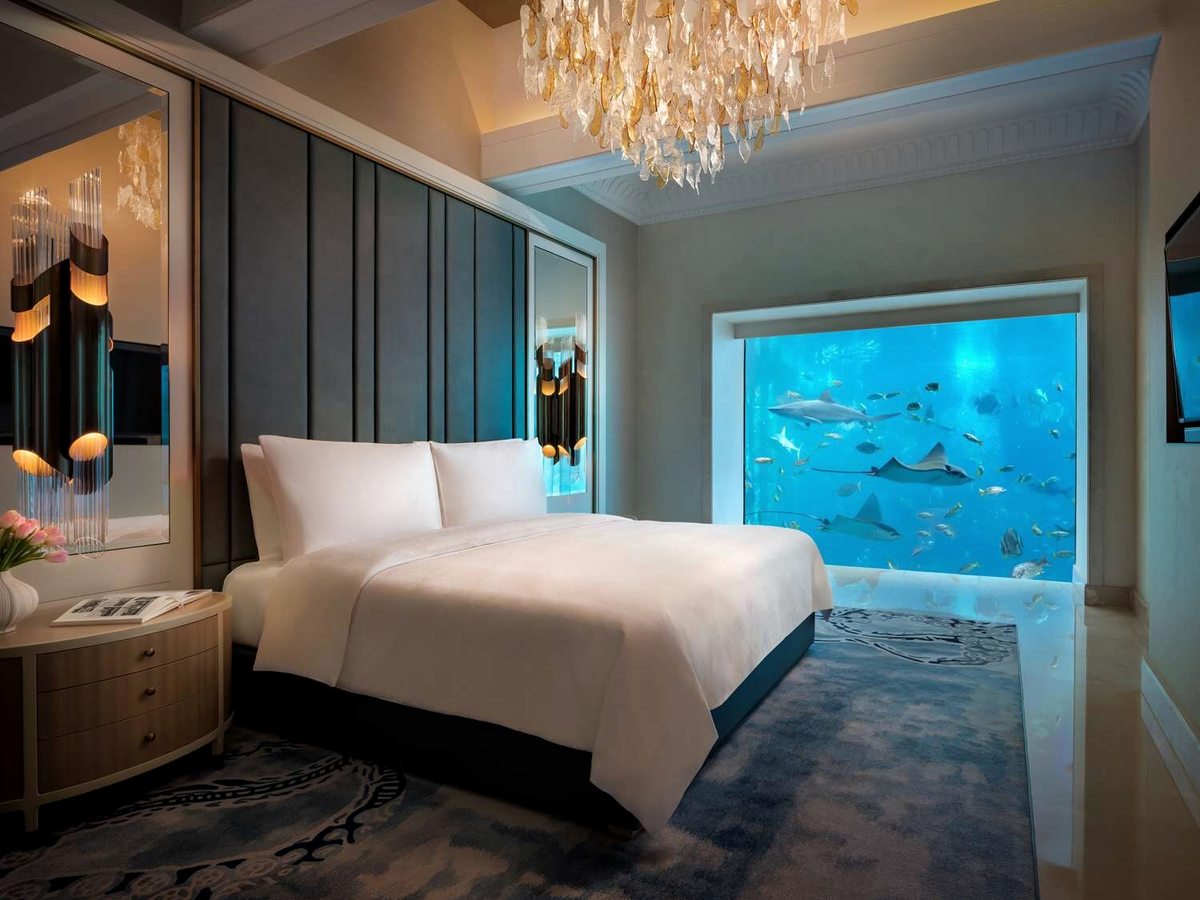 A suite bedroom with a king bed facing a floor-to-ceiling window that looks into an aquarium where a shark and rays swim, under a shell chandelier.