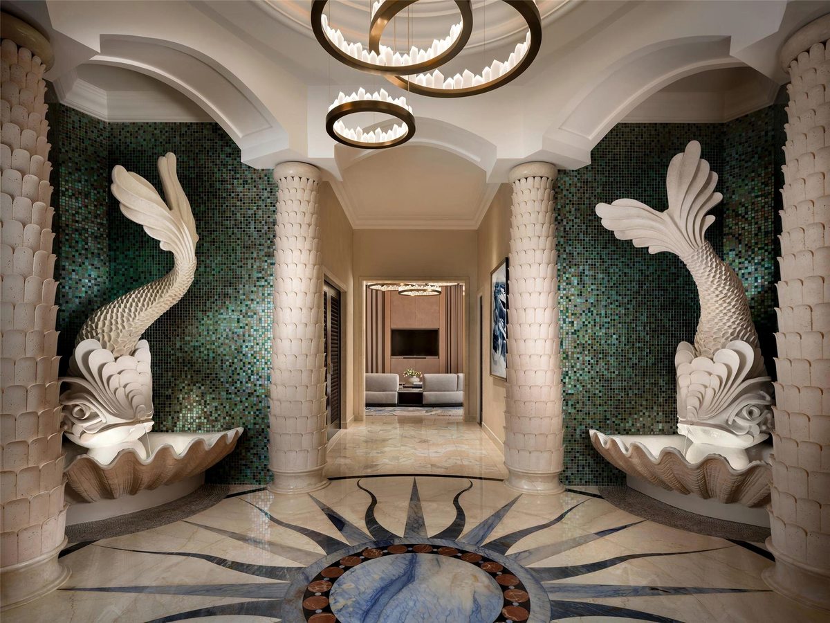 A grand lobby with two tall white fish sculptures spouting into shell fountains, green mosaic walls, stone columns and a compass motif set into the marble floor.