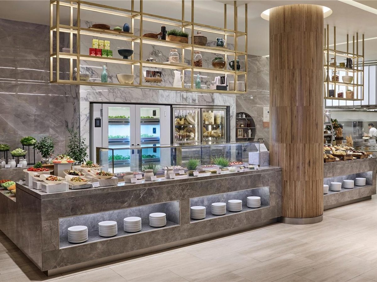 A long marble buffet counter laid with salads and cold dishes, stacked plates in recesses below, a wooden column and an open kitchen with a chef beyond.