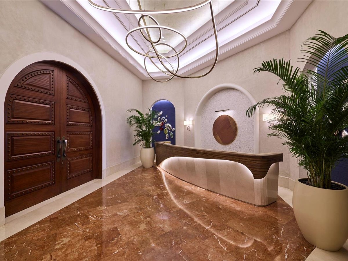 A curved timber reception desk beneath a looped ceiling light, beside tall arched wooden doors and potted palms on a polished marble floor.