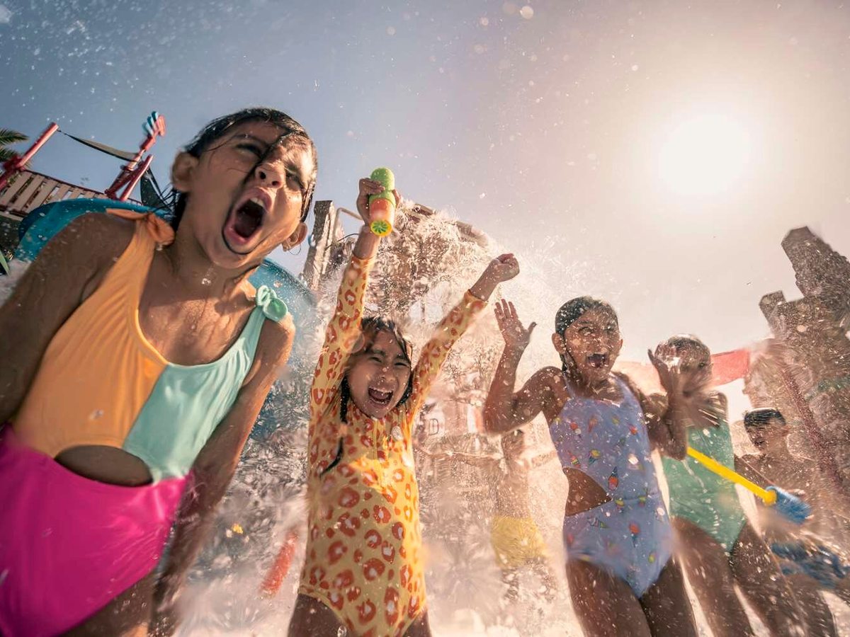 Young children in swimsuits laughing and playing under sprays of water in a splash area, backlit by bright sun.