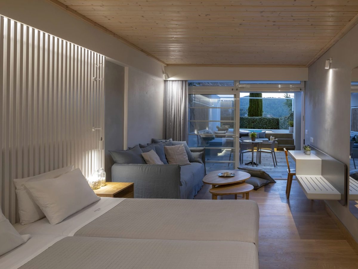 A suite with two single beds, a grey sofa and low tables, a slatted feature wall and sliding glass doors to a private terrace with a dining set.