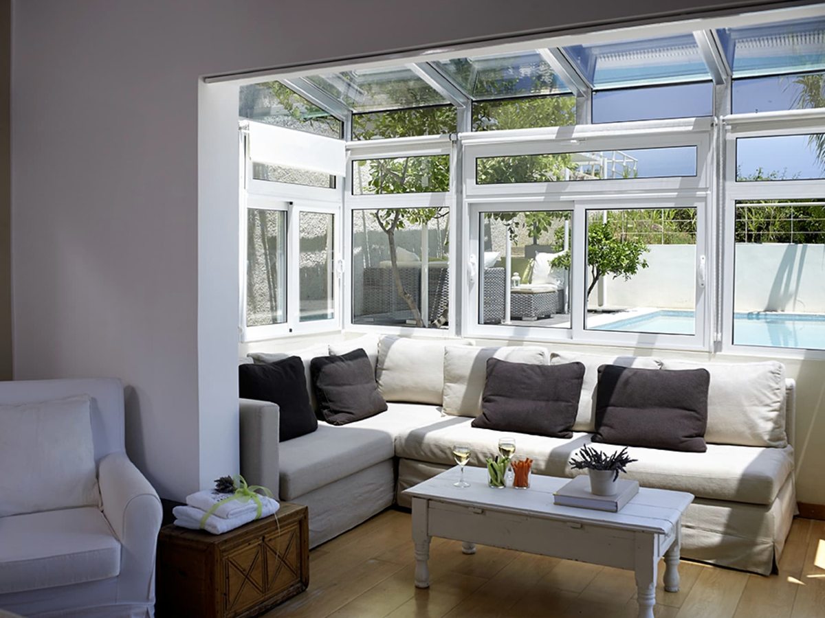A bright conservatory-style lounge with a glass roof, white sofas and a coffee table, with a private terrace and greenery beyond the windows.