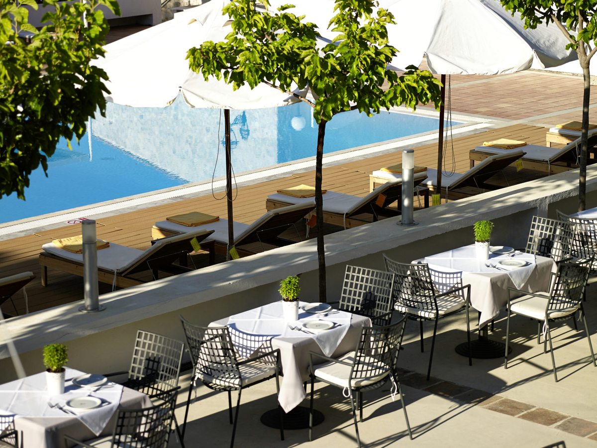 A raised dining terrace with white-clothed tables and metal chairs overlooking the swimming pool, its sun loungers and parasols below.