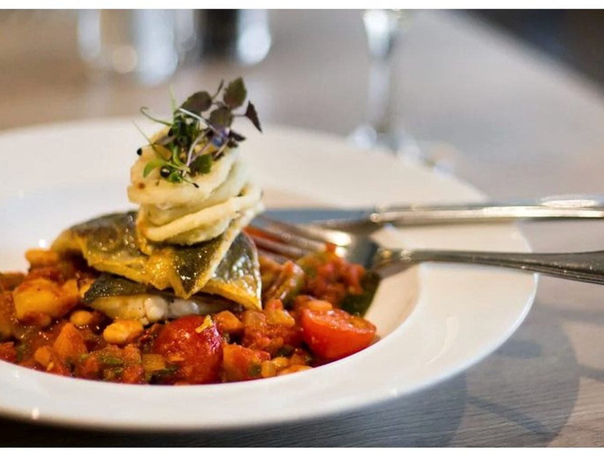 A fish main course on a tomato and vegetable ragout topped with microgreens, served in a shallow white bowl on a wooden table.