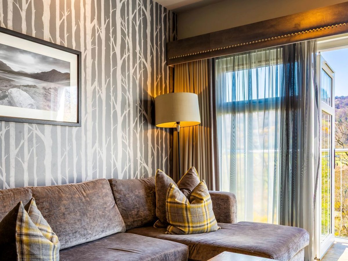 A room's seating corner with a brown velvet sofa beside an open balcony door looking onto autumn woodland, under birch-tree wallpaper.