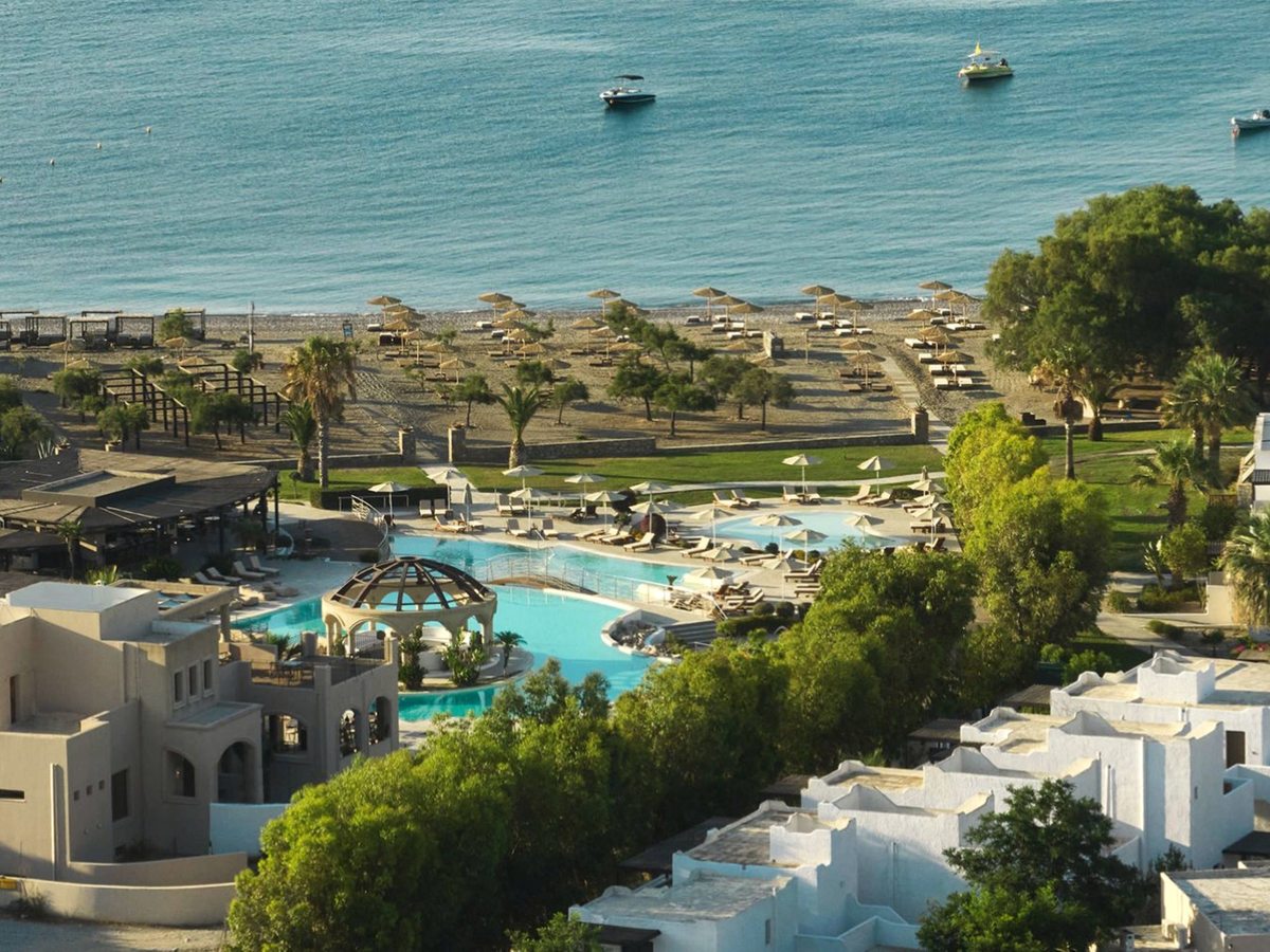 Elevated view of the resort pools and white village-style buildings, with the beach and calm sea just beyond.