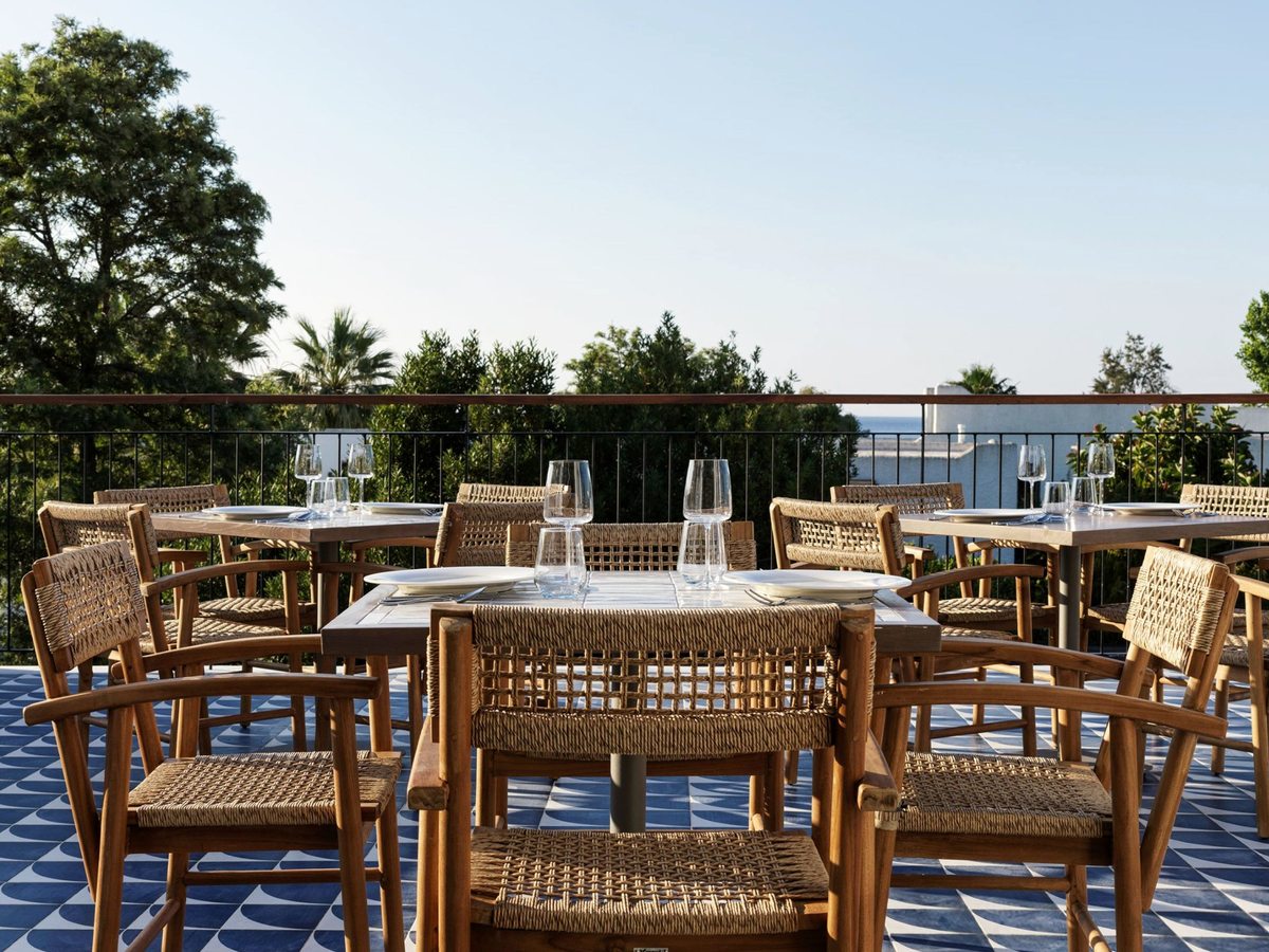 Outdoor restaurant terrace with woven chairs and set tables on a blue-patterned floor, the sea beyond.