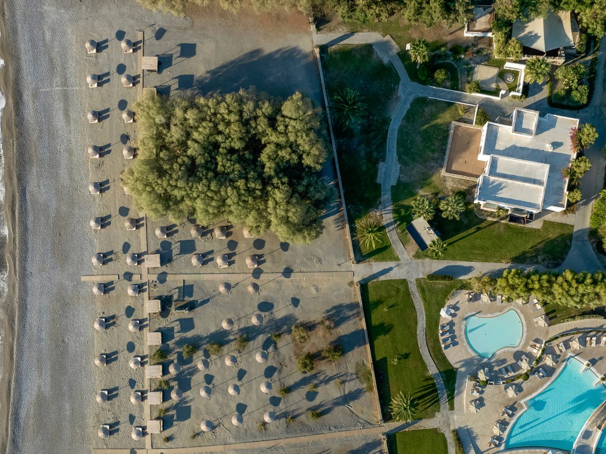 Overhead view of a beach lined with straw parasols, with the resort's pools and lawns alongside.