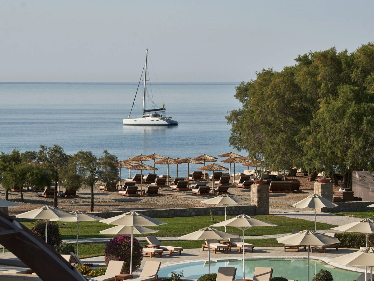 View over garden parasols and beach loungers to a calm sea with a moored catamaran offshore.
