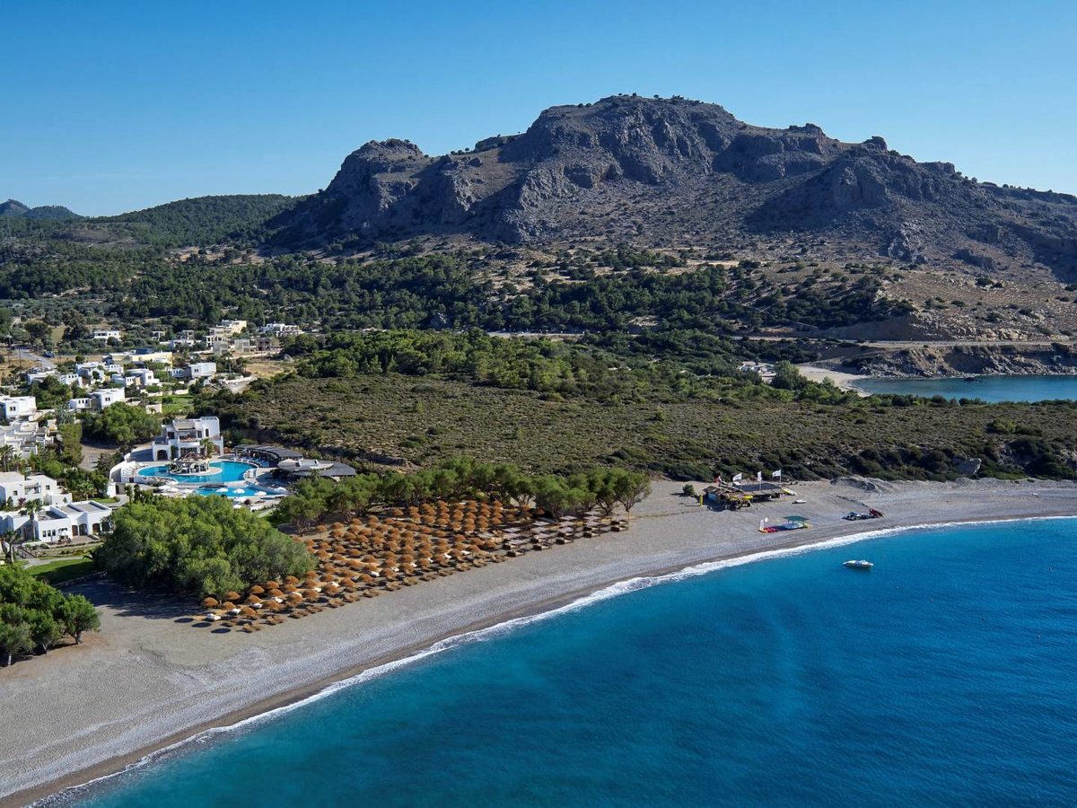 Aerial view of the resort's white buildings and pool beside a long beach, with a rocky headland rising behind.