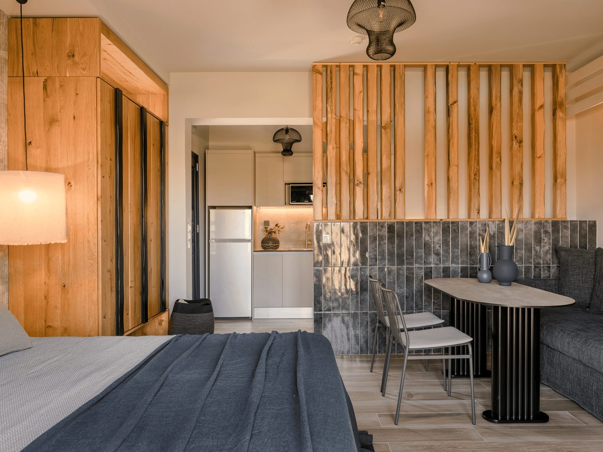 Open-plan studio with a double bed, a grey sofa and a dining table, and a kitchenette with a full-size fridge and microwave behind a timber-slat divider.