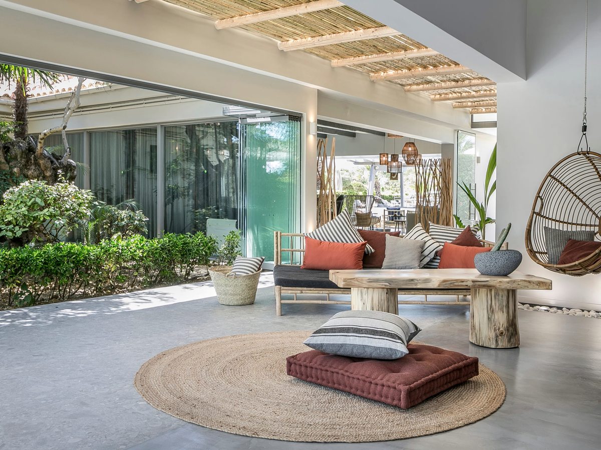 Open-sided covered terrace with a cushioned daybed, orange and striped cushions, a hanging rattan chair and a rustic tree-trunk table on a round woven rug.