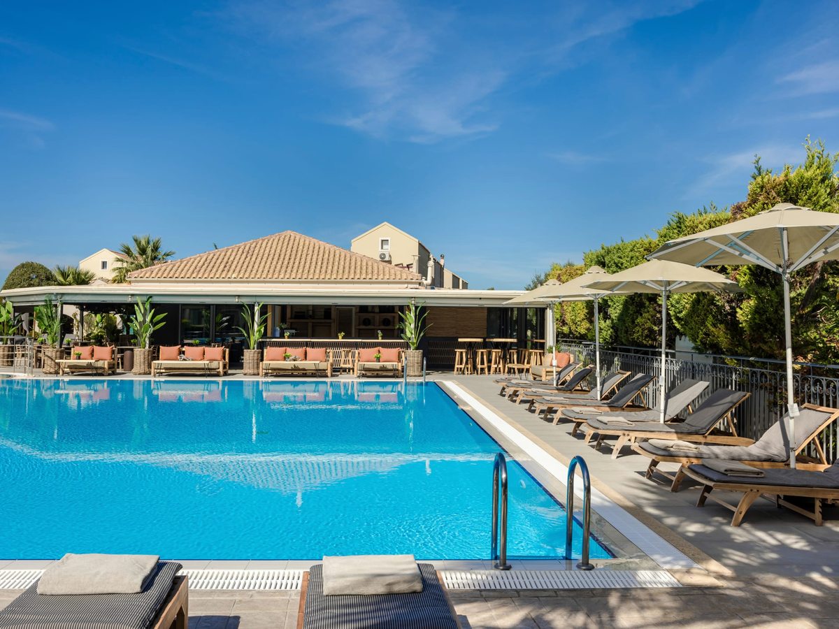 Large rectangular pool with wooden sun loungers and white parasols along the tiled surround and the low resort buildings behind.