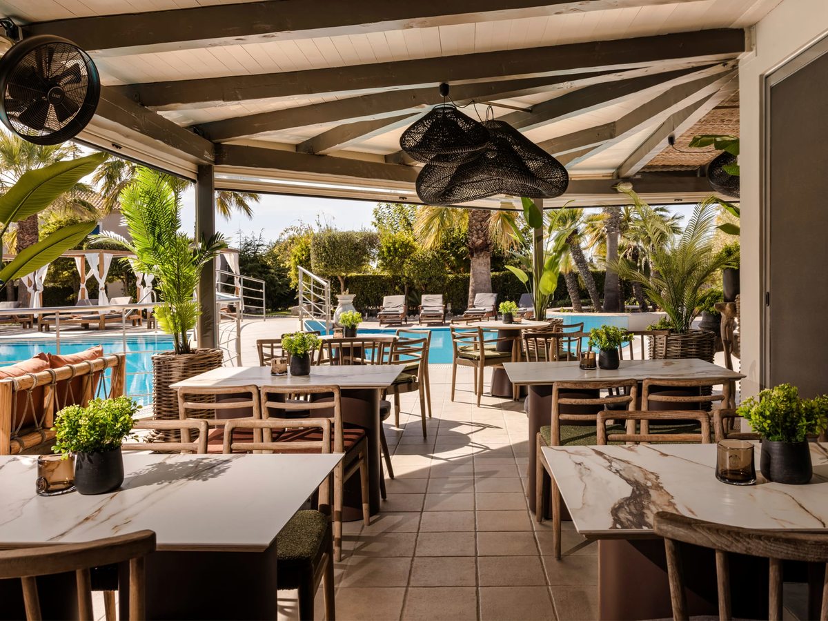 Covered poolside terrace with marble-top tables, wooden chairs and ceiling fans looking out over the pool and its loungers.