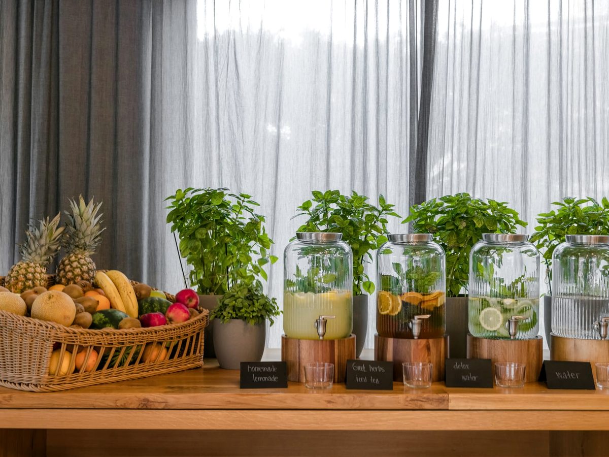 Buffet counter set with glass jars of infused water and juice, tumblers and baskets of pineapples, bananas and oranges in front of curtained windows.