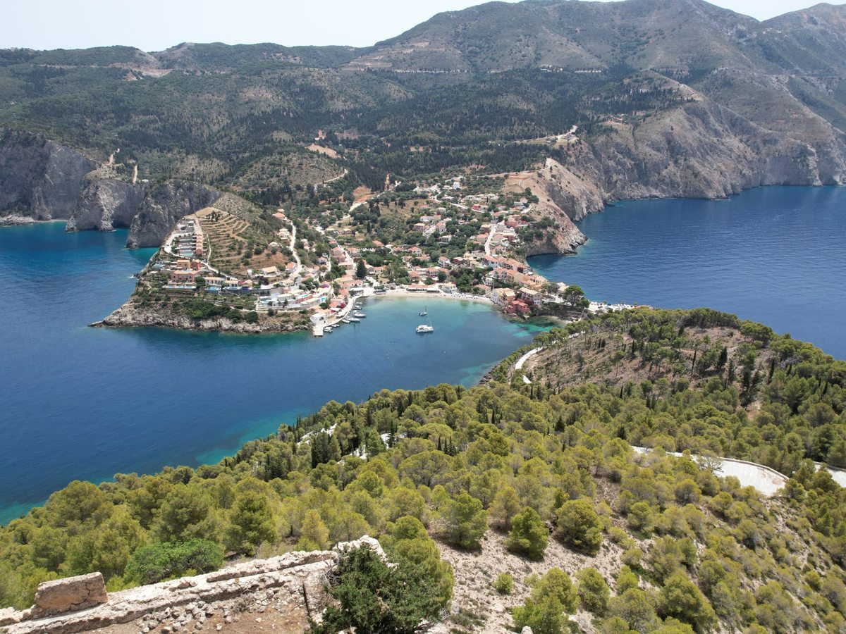 Elevated view of Assos village on a wooded peninsula, with a sheltered bay on either side and mountains beyond.