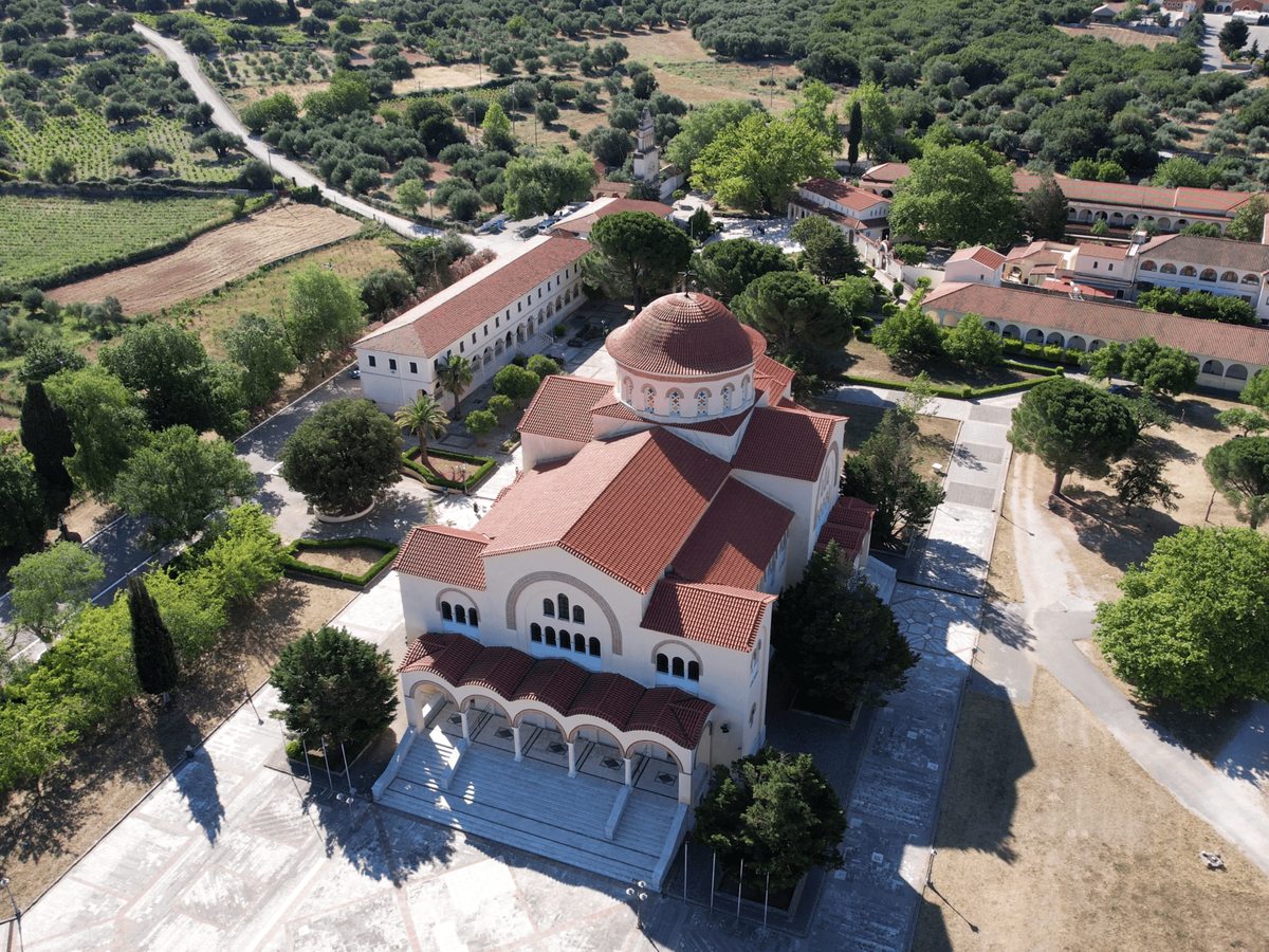 Aerial view of a white church with a red-tiled roof and bell tower set among fields and vineyards in the countryside.
