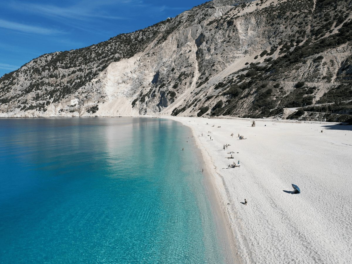 Crescent white-pebble beach backed by tall grey limestone cliffs, with turquoise shallows deepening to blue and a few beachgoers on the sand.