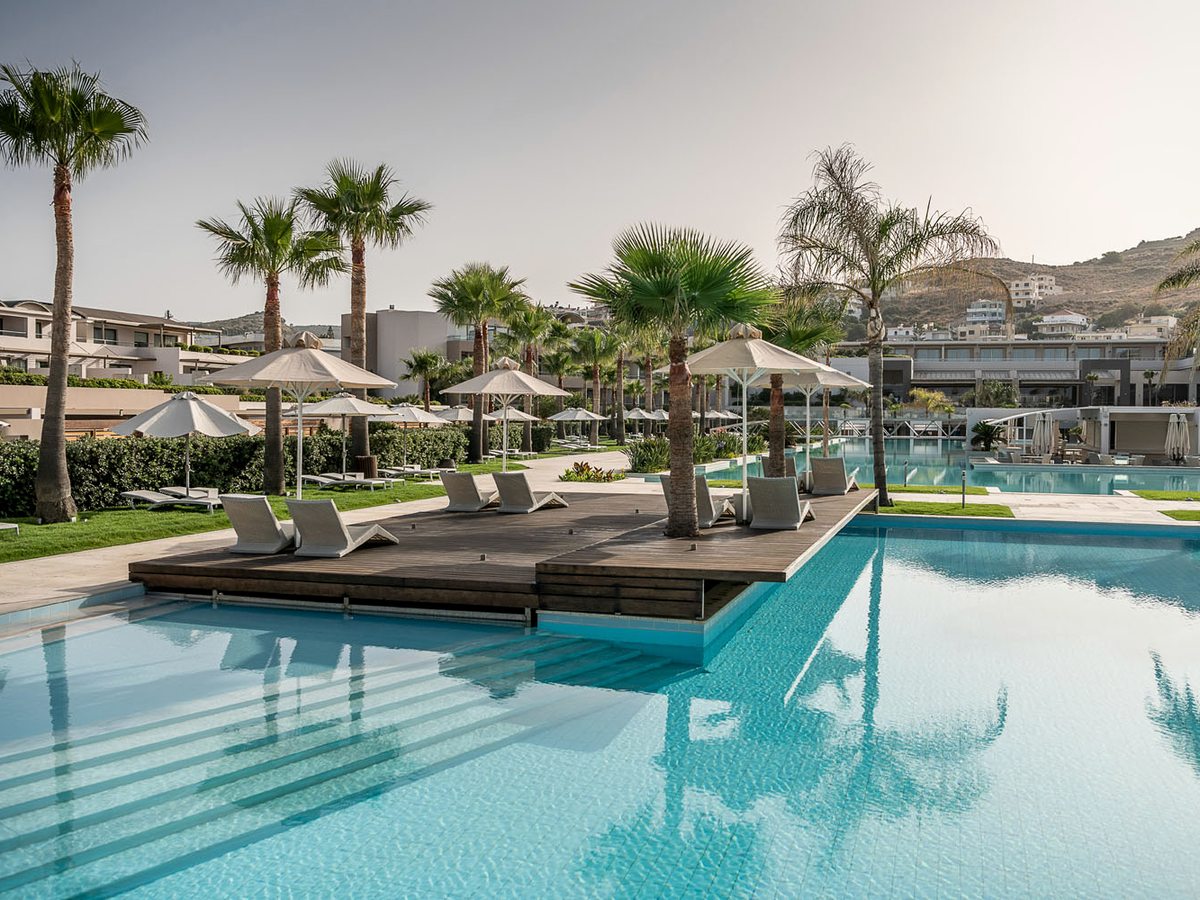 An outdoor pool with a raised wooden sun deck and loungers between two stretches of water, palms and hills behind.
