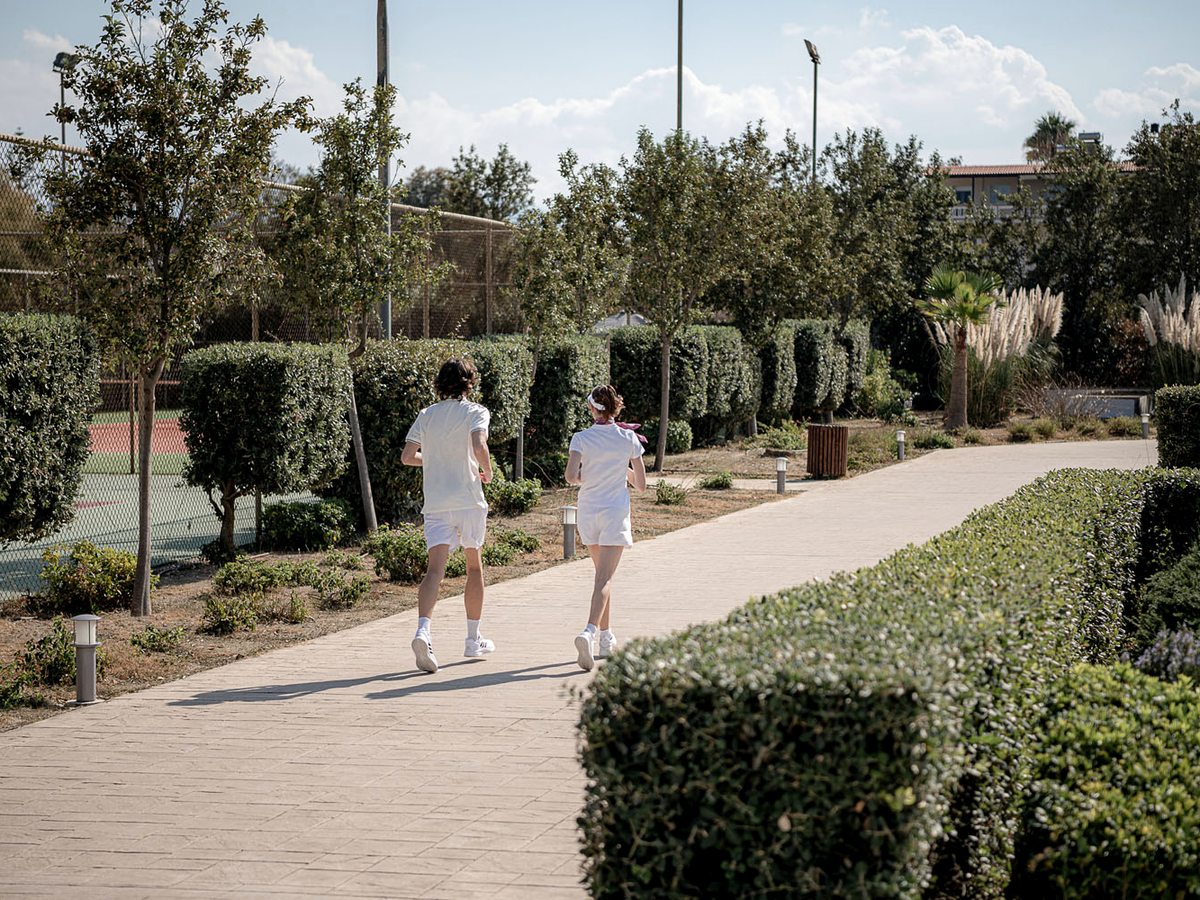 A paved garden path between clipped hedges and olive trees, with two people walking and low buildings beyond.
